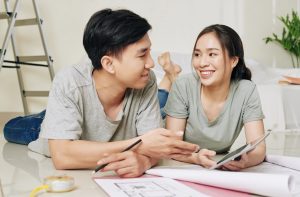 Beautiful young Vietnamese couple with tablet computer lying on the floor and discussing apartment plan