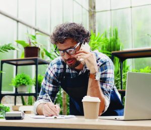 Happy Caucasian businessman talking on cellphone while standing in apron in small floral center and writing down order details. Joyful male florist calling on smartphone at work. Own business concept